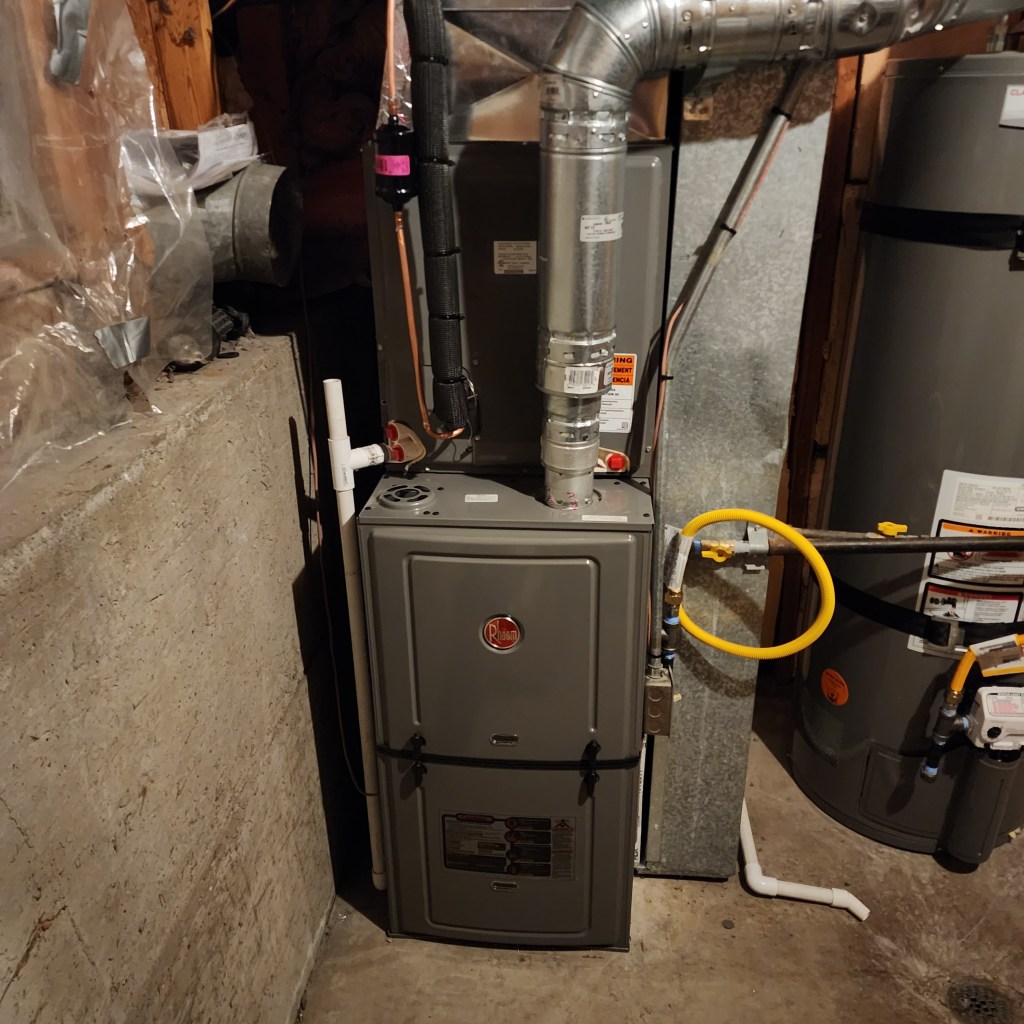 Photo of a Rheem gas furnace in the basement of a home. A metal exhaust flue is coming out the top and a yellow flexible gas line is attached to the side.